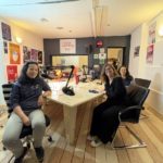 Florence, Emmanuelle et Céline autour de la table en bois du studio. Sur la table on voit 4 micros. Il y a des affiches sur les murs du studio.