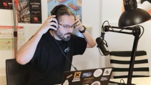 Fred, habillé d'un t-shirt, avec les mains sur le casque, devant un micro.